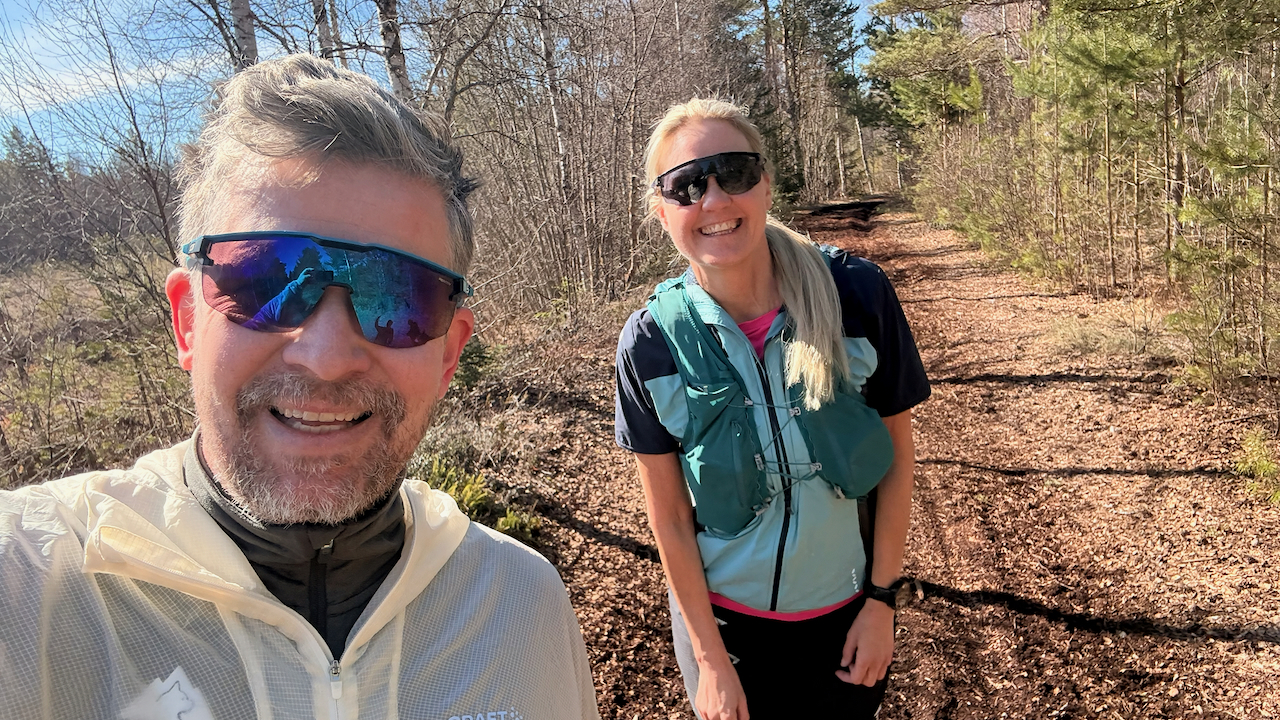 Marcus and Mimmi wearing athletic gear and sunglasses are smiling while trail running on a sunny day in the woods.