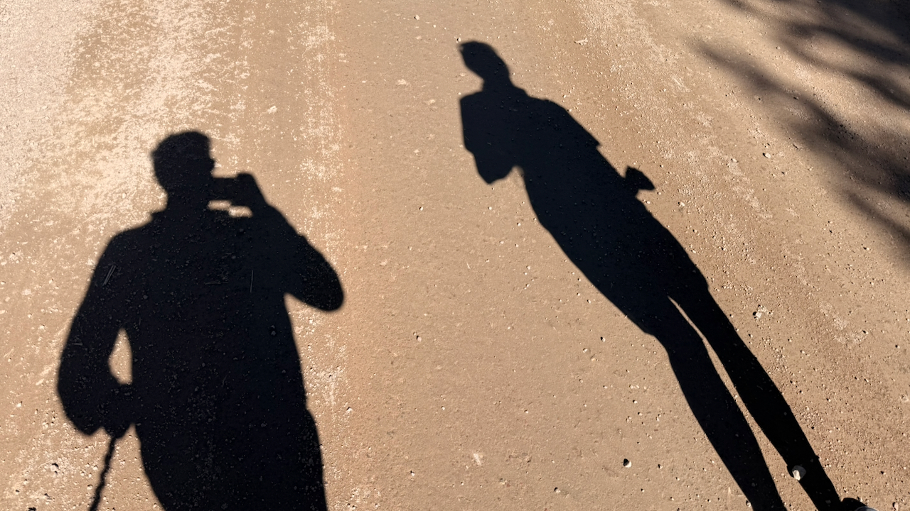 The long, morning shadows of two people are cast onto a dusty dirt path. One shadow appears to be taking a photo while the other person is walking.